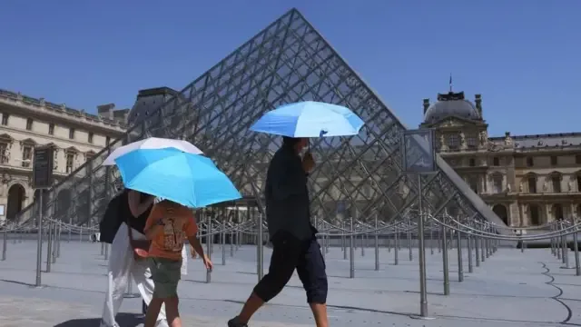 Tres turistas se protegen del sol con sombrillas mientras caminan frente a la Pirámide del Museo del Louvre.