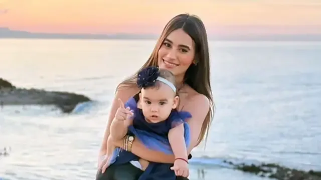 Una mujer de cabello largo y oscuro sonríe mientras sostiene a una niña pequeña vestida con un vestido en la playa, con el atardecer de fondo.