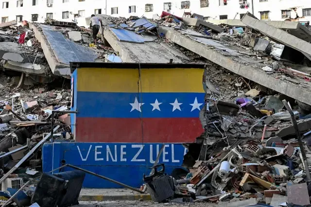 Un muro en el que estaba pintada la bandera de Venezuela yace roto frente a un edificio desplomado por los terremotos en La Guaira, 26 de junio 2026
