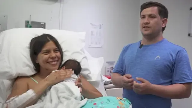Dayana Patiño sonríe mientras sostiene a su hijo Juan David contra su pecho desde una cama de hospital. Su esposo, Gerson, está de pie junto a ellos vistiendo una camiseta azul claro.