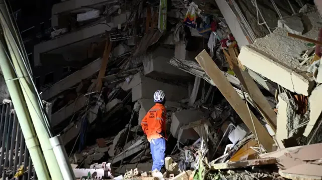 Un socorrista con una chaqueta naranja brillante mira fijamente un edificio derrumbado, con un enorme montículo de escombros, bloques de hormigón y acero retorcido a la vista.