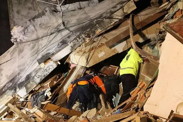 Equipos de rescate buscan un edificio derrumbado: uno con una chaqueta naranja de alta visibilidad, otro con pantalones negros y otro con una chaqueta verde de alta visibilidad.