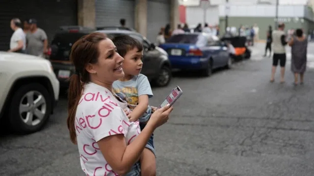 Una mujer con cara de angustia carga a un niño pequeño con una mano mientras sostiene su celular con la otra mano en una calle de Caracas, luego del doble terremoto del 24 de junio.
