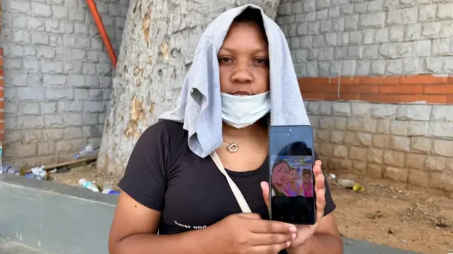 Una mujer con una camiseta gris que cubre su cabeza sostiene un teléfono móvil para mostrar una foto suya con sus dos hijas adolescentes.
