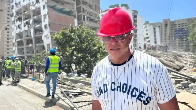 Un hombre con un casco rojo y gafas de sol está parado en una calle frente a los escombros de un edificio derrumbado, y en las inmediaciones hay funcionarios con chalecos de alta visibilidad.