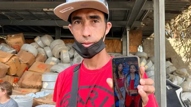 Un hombre con una gorra de béisbol y una camiseta roja muestra una foto de su esposa e hija en un teléfono móvil que sostiene en alto con la mano izquierda.