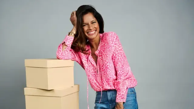 Dilly llevaba una camisa rosa estampada de manga larga, apoyada en una pila de cajas de color beige en primer plano, con una mano apoyada en un costado de la cabeza. Se ven varias pulseras, anillos y un collar. El fondo es una pared lisa de color gris claro.