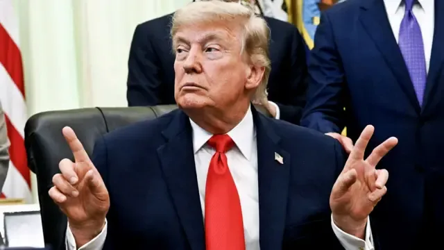 El presidente de Estados Unidos, Donald Trump, señalando con el dedo en el Despacho Oval de la Casa Blanca, con una bandera estadounidense y hombres detrás de él.